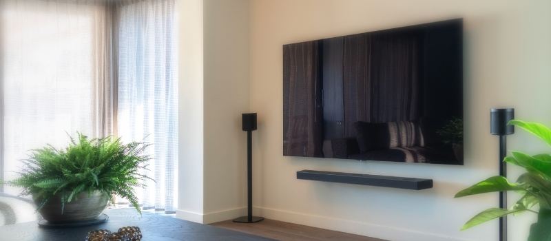 Soggiorno con tende bianche a sinistra e televisore con soundbar e due altoparlanti separati su supporti a destra