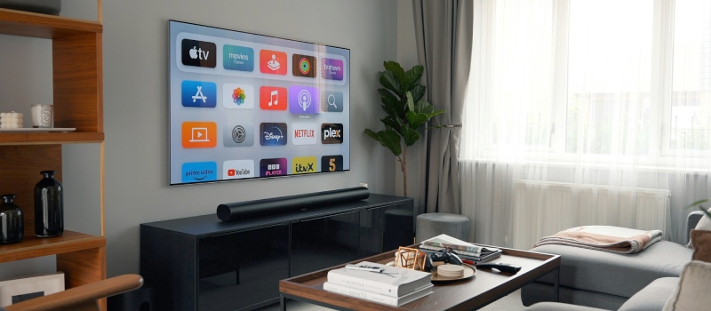 una televisione appesa alla parete con una soundbar su un mobile TV nero, a sinistra della TV un armadio in legno e a destra una pianta verde