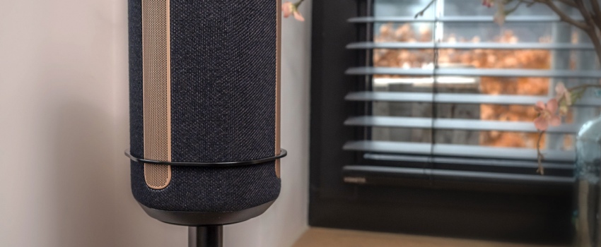 Primo piano di un altoparlante nero su un supporto Vebos con staffa, collocato in un interno moderno vicino a una finestra con tende veneziane.